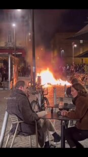 French couple enjoy glass of red wine as street burns behind them